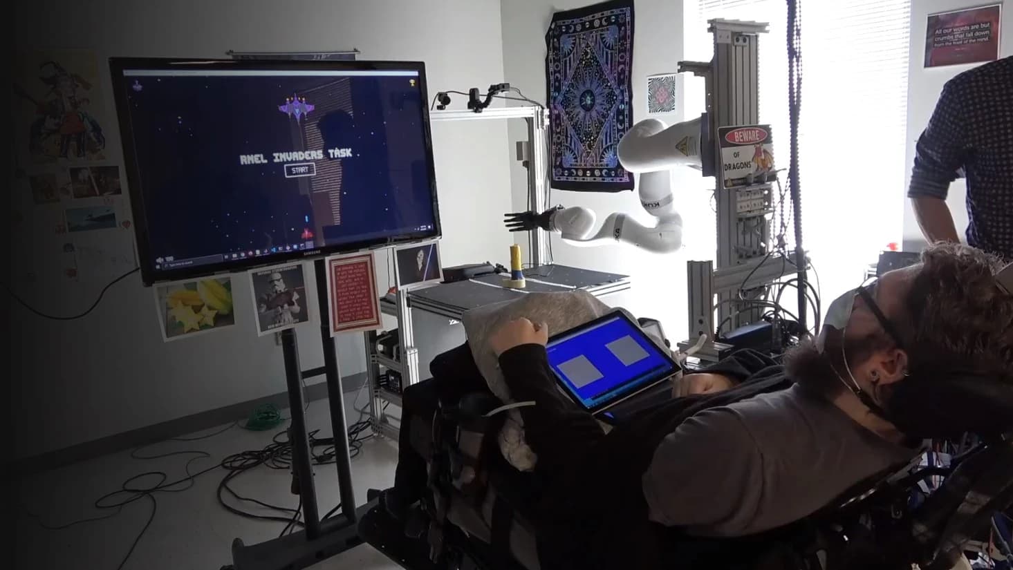 Research participant in the Rehab Neural Engineering Labs during a gamified intracortical BCI calibration session, with the task displayed on a monitor and a tablet controller nearby